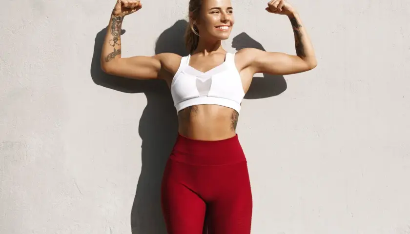 Uśmiechnięta kobieta w sportowym stroju pokazuje swoje silne ramiona, efekt ciężkich treningów i pompki na biceps.