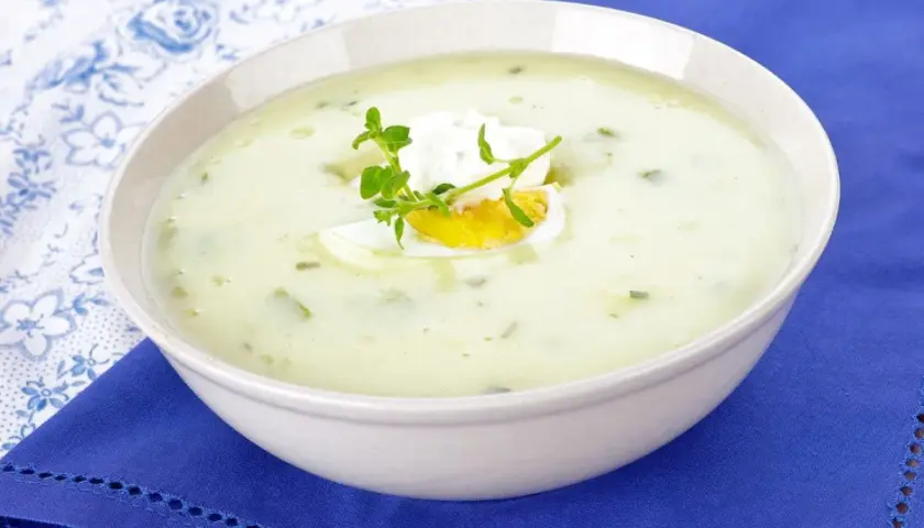 Kremowa zupa z sałaty z jajkiem i ziołami, podana w białej misce na niebieskim obrusie.
