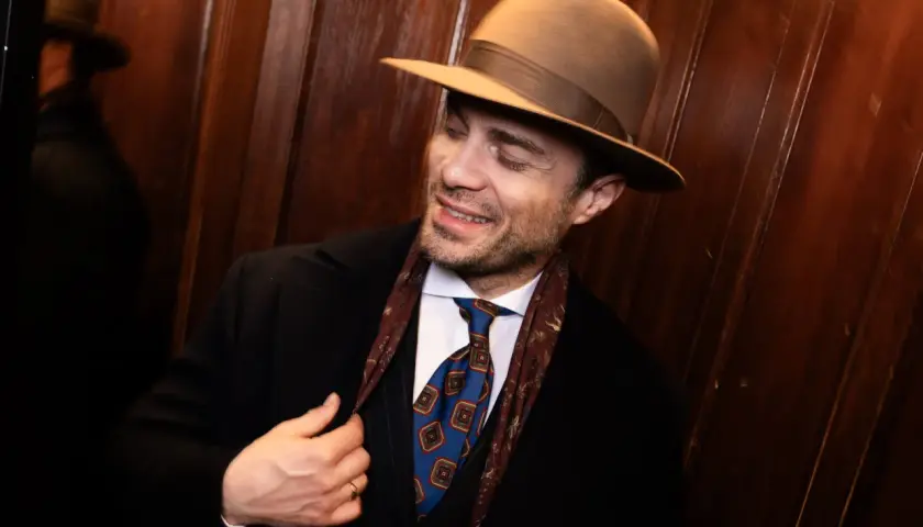 Elegancki mężczyzna w brązowym kapeluszu fedora, z krawatem i apaszką, poprawia marynarkę.