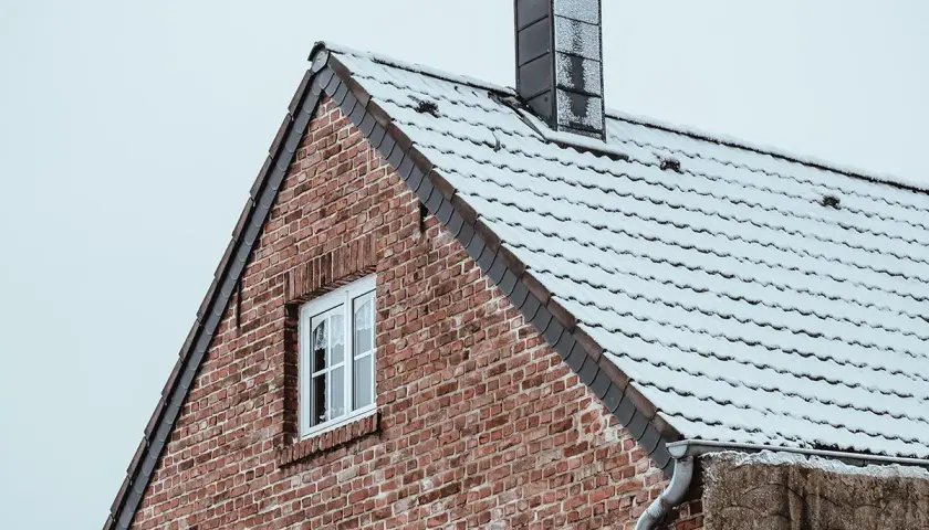 Ett tegeltak med snö och en mörk rökkanal skorsten mot en grå himmel.