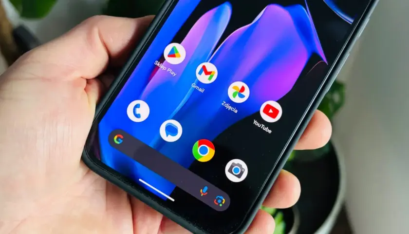 Dłoń trzyma smartfon z kolorową tapetą i ikonami aplikacji, w tym Sklep Play, Gmail, Zdjęcia i YouTube. Wygląda jak nowy **android 15**.