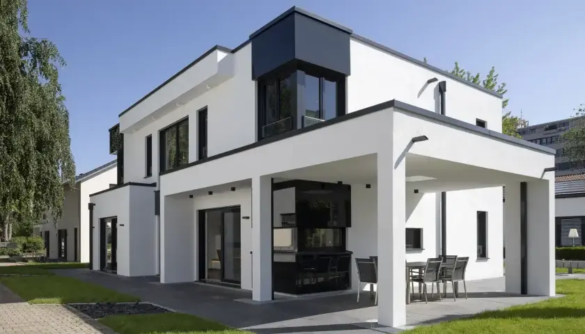 Ein modernes Haus mit Flachdach, weißen Wänden und schwarzen Fensterrahmen. Eine überdachte Terrasse mit Esstisch lädt zum Verweilen ein.