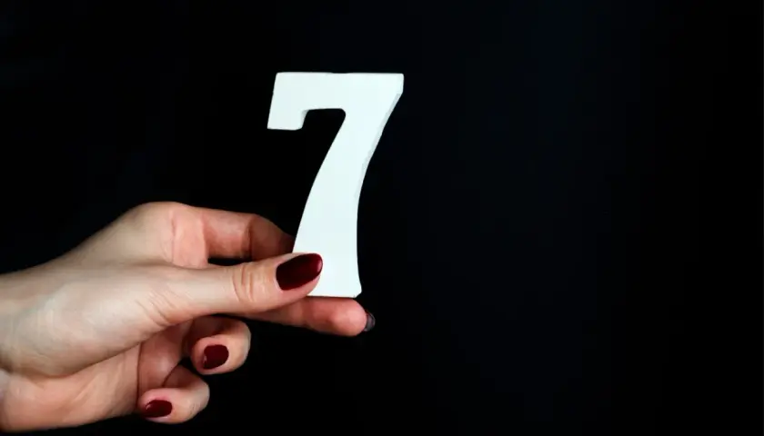 Dłoń z ciemnoczerwonym lakierem trzyma białą cyfrę 7, symbolizującą numerologiczną 7.