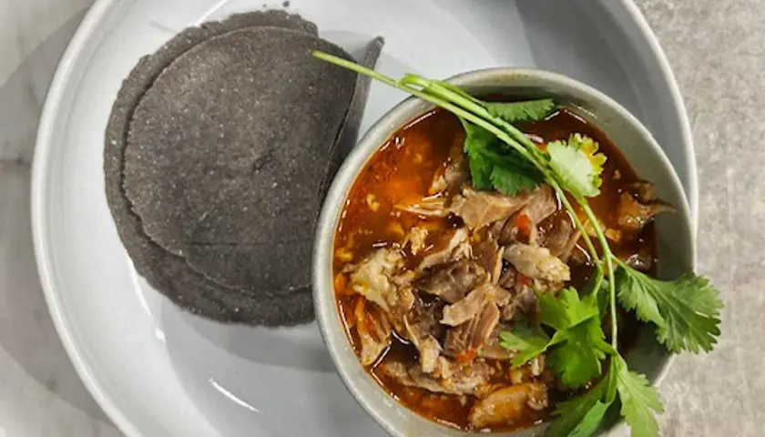 En skål med mustig el birria, toppad med koriander, serveras med mörka majstortillas på en vit tallrik.
