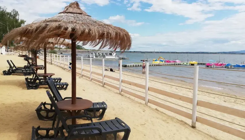 Plaża nad otmuchowskim jeziorem, leżaki pod strzechą, w oddali kolorowe rowerki wodne i statek wycieczkowy.