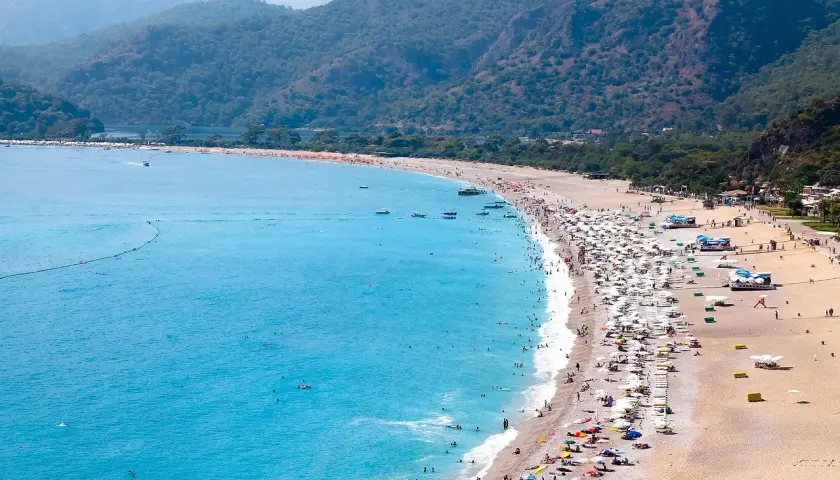 Tłum ludzi odpoczywa na piaszczystej plaży, kąpiąc się w turkusowym morzu. Widok na riwierę egejską z górzystym, zalesionym tłem.