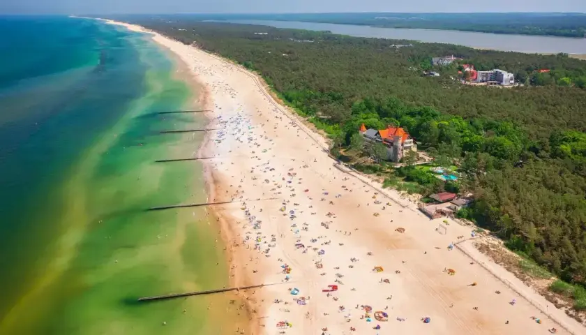 Słoneczna plaża nad Jeziorem Łebsko, pełna wypoczywających ludzi i zielony las. Atrakcje turystyczne w zasięgu wzroku.