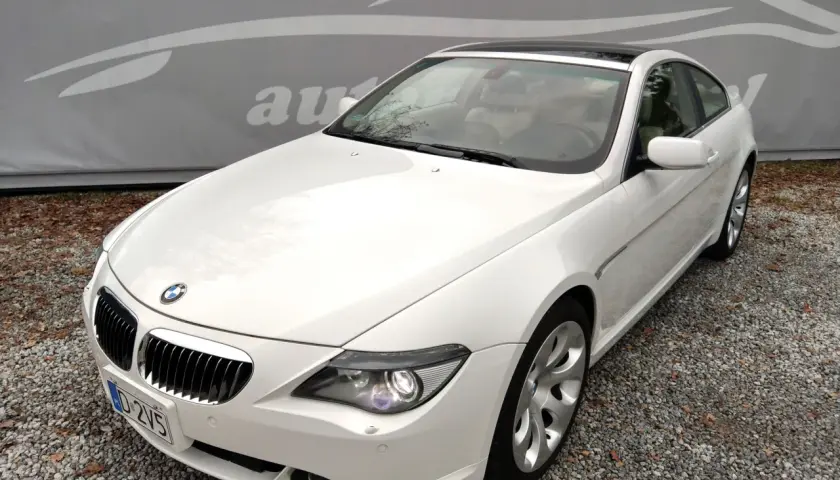 Białe BMW E63 coupe z jasnym wnętrzem, zaparkowane na żwirze, z logo autokomisu w tle.