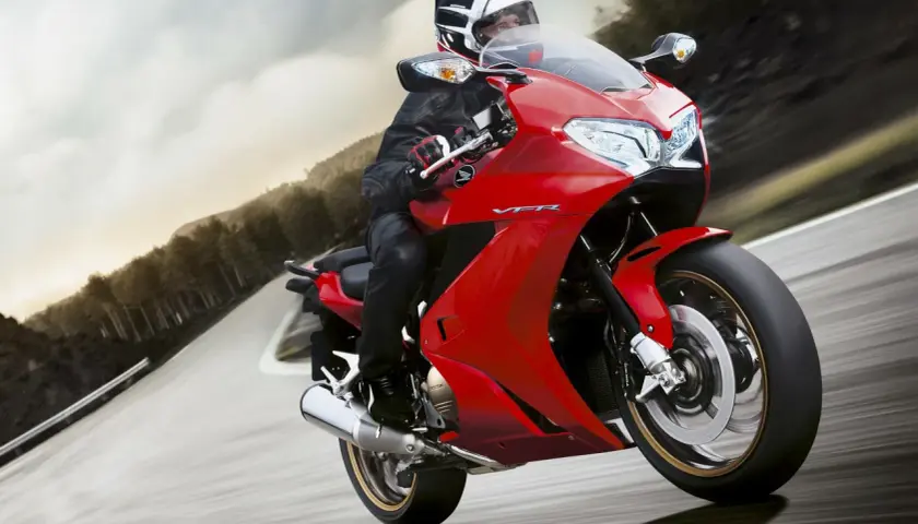 Motociclista en moto roja deportiva, con casco y equipo de cuero, inclinándose en una curva. El carenado de protección de la moto es aerodinámico y brillante.
