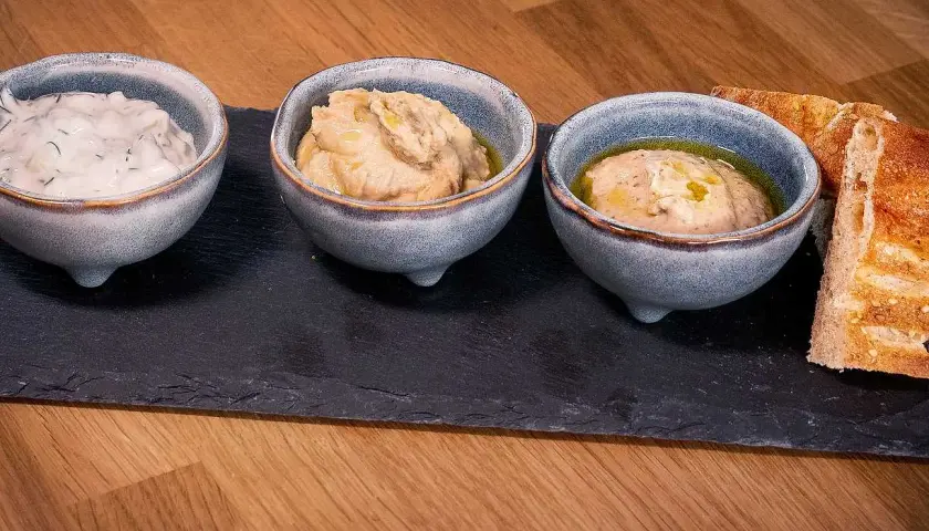 Tre skålar med meze recept: en med vit sås och dill, två med hummusliknande röror. Serveras med rostat bröd på en svart skifferplatta.