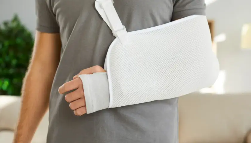 Osoba w szarym t-shircie nosi biały temblak na rękę. Czy trzeba nosić cały czas temblak? To pytanie zadaje sobie wielu po urazie.