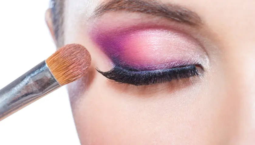 Pędzel do makijażu tworzy wyrazisty **cut crease** na powiece, podkreślając oko intensywnym fioletem i różem.