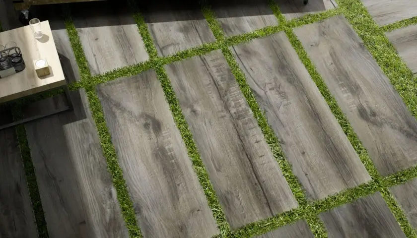 Pavimento posa a secco su erba, con doghe effetto legno grigio alternate a strisce di prato verde. Un tavolino da caffè completa la scena.
