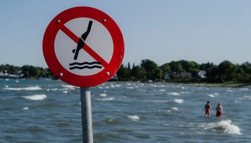 Znak zakazu kąpieli nad morzem. Dwie osoby w wodzie ignorują ostrzeżenie.