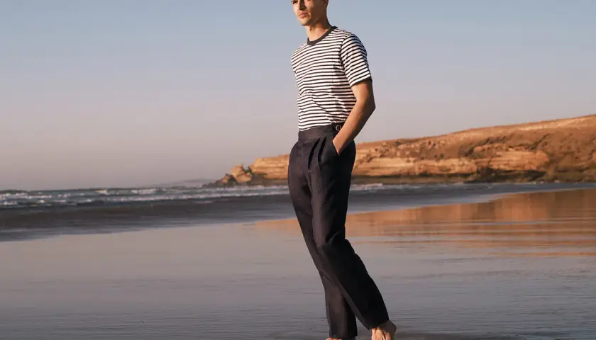 Mężczyzna w granatowych chinosach i pasiastym T-shircie spaceruje po plaży. Chinosy co to? To uniwersalne spodnie na każdą okazję.