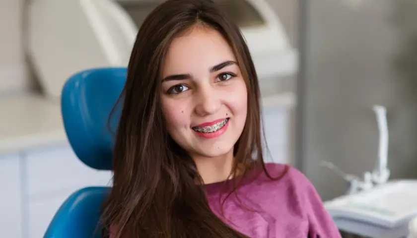 Uśmiechnięta nastolatka z kolorowym aparatem ruchomym na zębach siedzi na niebieskim fotelu dentystycznym.
