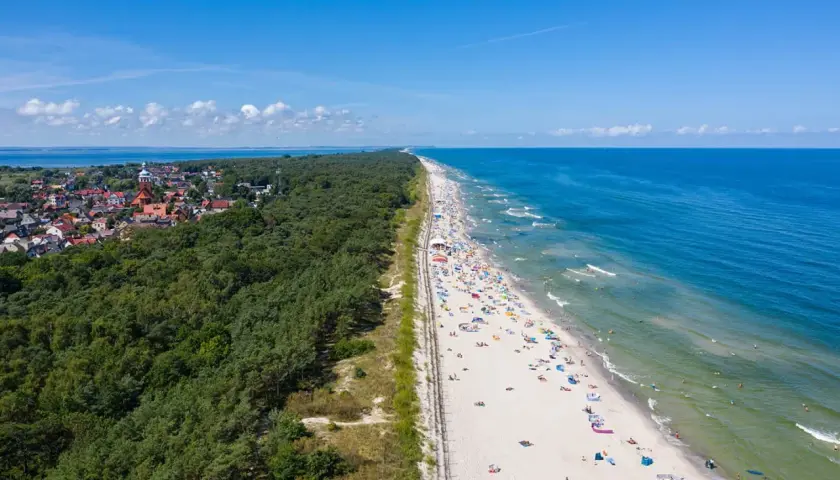Breiter Sandstrand mit vielen Badegästen, gesäumt von einem dichten Kiefernwald und einer kleinen Stadt. Ein echter Geheimtipp an der polnischen Ostsee.