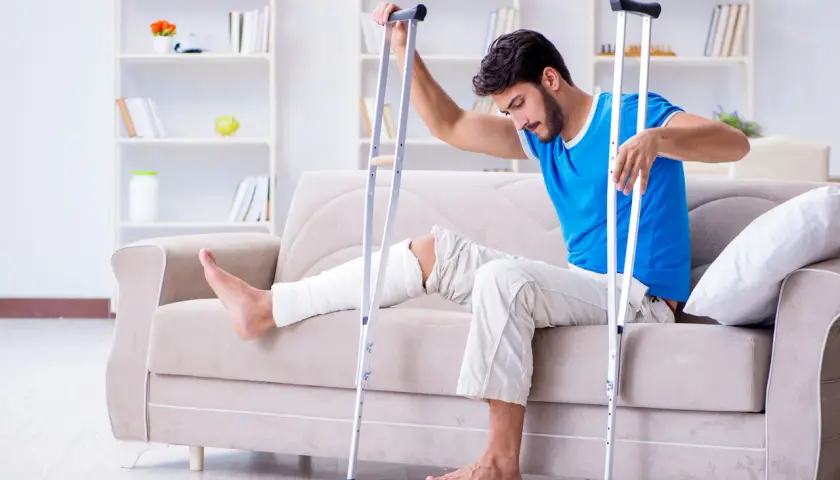 Mężczyzna z zabandażowaną nogą siedzi na kanapie, opierając się o kule. Czeka na rehabilitację po kontuzji, która uniemożliwia mu chodzenie o kulach.