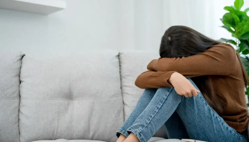 Kobieta siedzi skulona na kanapie, przytulając kolana. Jej twarz jest ukryta, co może symbolizować depresję lękową i poczucie osamotnienia.