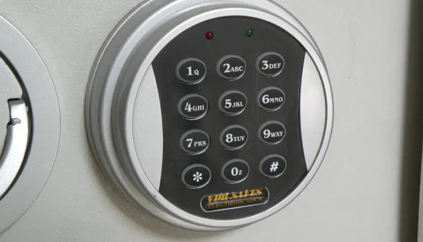 Elektroniczna klawiatura szyfrowa sejfu z przyciskami cyfrowymi i symbolami. W tle widoczne są metalowe tuleje mocujące.