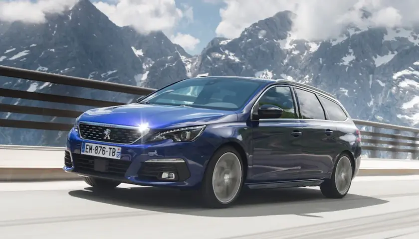 Niebieski Peugeot 308 T9 kombi jedzie po górskiej drodze, z ośnieżonymi szczytami w tle.
