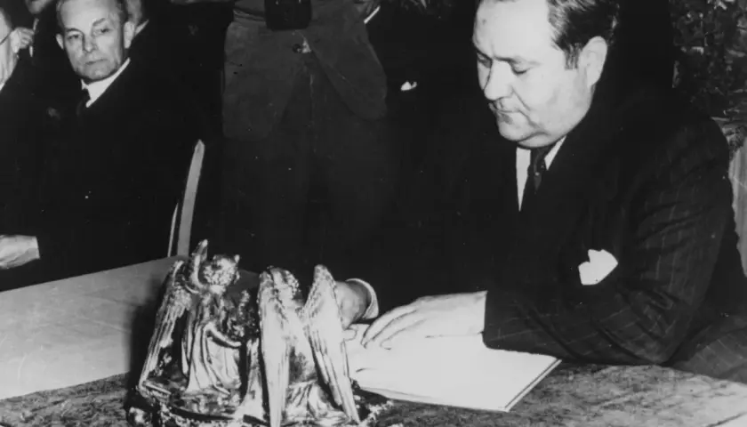 Carlo Schmid sitzt an einem Tisch und blickt auf ein Buch. Ein Fotograf hält eine Kamera.