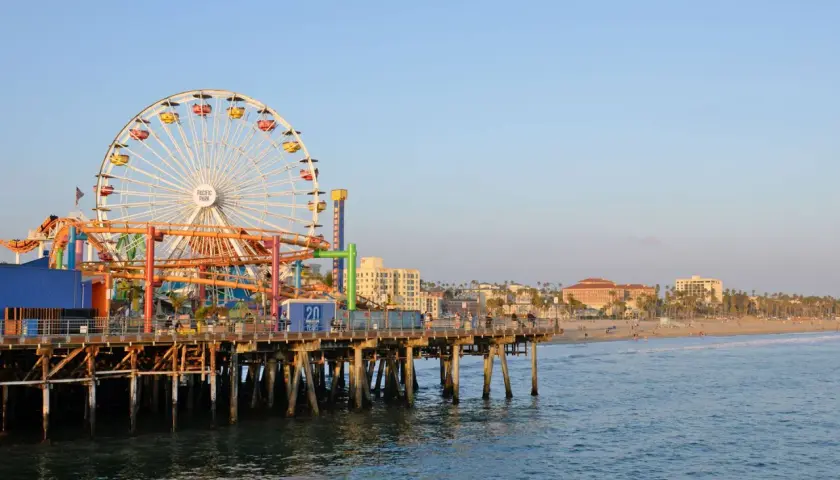 Der Santa Monica Pier mit seinem Riesenrad und Achterbahn ragt ins Meer. Im Hintergrund erstreckt sich der sonnige los angeles strand mit Palmen und Gebäuden.