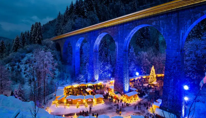 Magischer weihnachtsmarkt deutschland geheimtipp unter beleuchteter Brücke. Ein Zug fährt vorbei, während Besucher Buden und einen Weihnachtsbaum bestaunen.