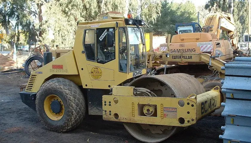 Żółty walec drogowy BOMAG BW 213D-3, obok koparka Samsung Caterpillar. Szerokość koparki nie jest widoczna, ale walec jest imponujący.