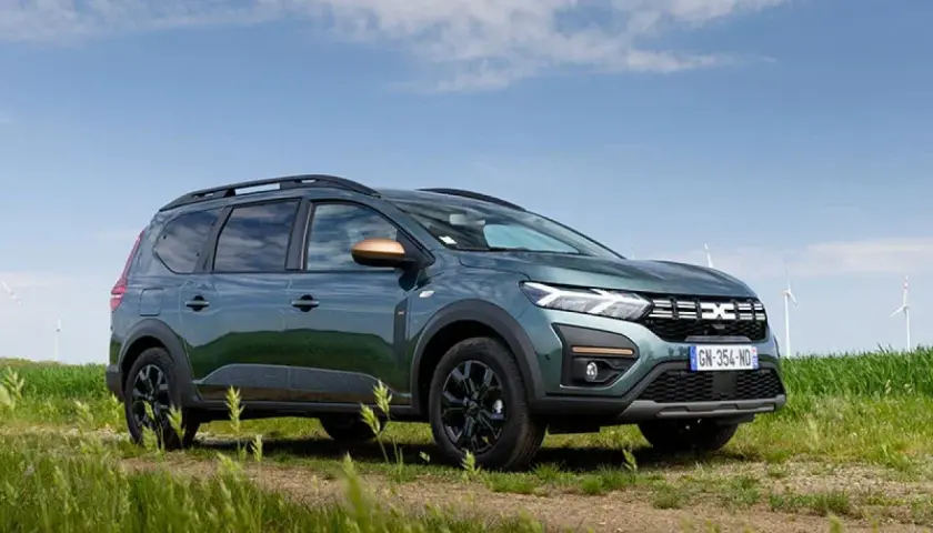 Un'auto familiare verde scuro, con barre sul tetto e dettagli color rame, parcheggiata su un sentiero sterrato in un campo con mulini a vento sullo sfondo.