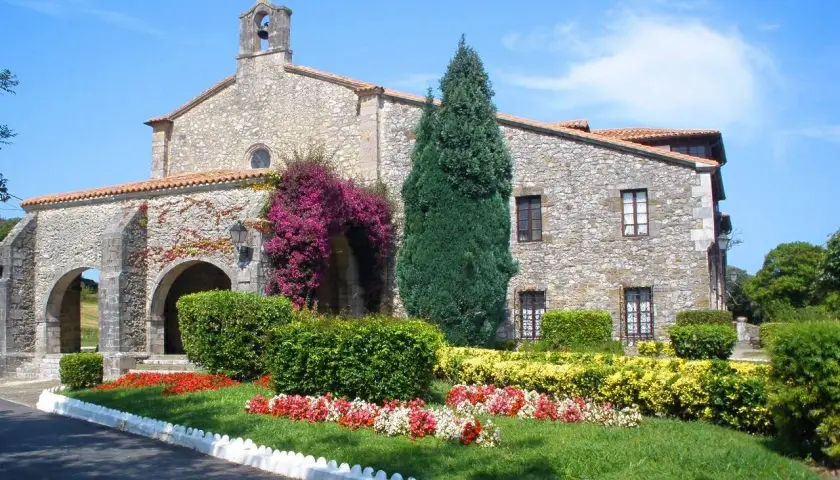 El **santuario de la Barquera**, con su fachada de piedra y arcos, se adorna con vibrantes buganvillas y un jardín florido bajo un cielo azul.