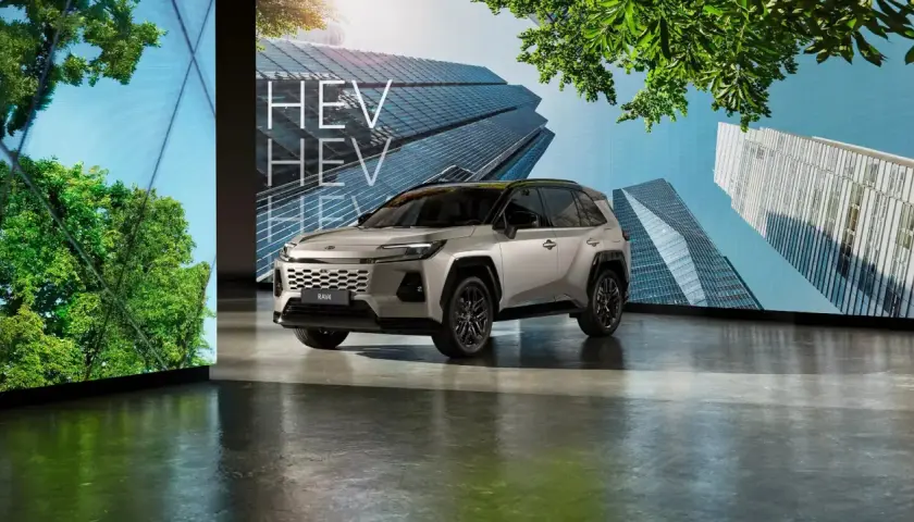 Srebrny SUV RAV4 HEV stoi przed ekranem z nowoczesnymi budynkami i zielenią.