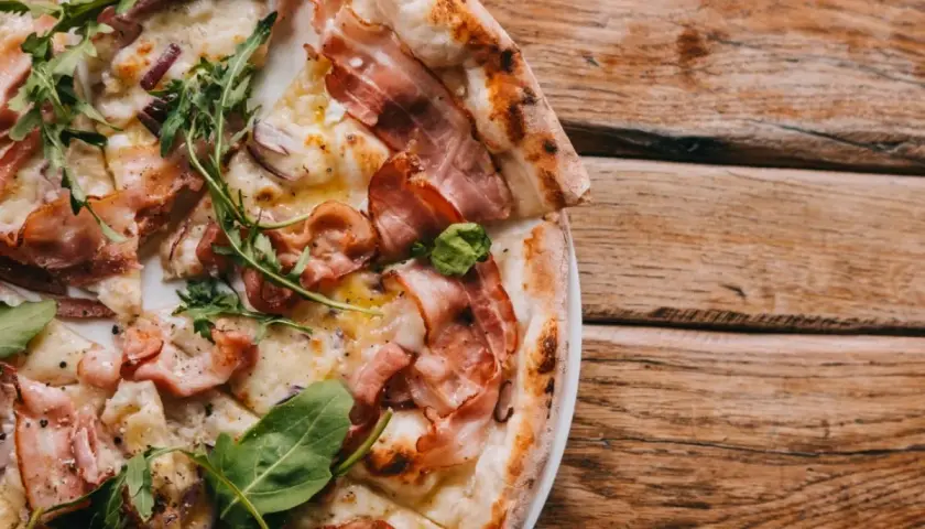 Pyszna pizza carbonara z chrupiącym boczkiem i rukolą, podana na drewnianym stole.