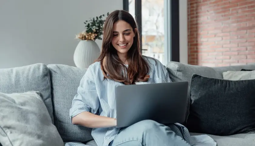Uśmiechnięta kobieta na kanapie, korzystająca z laptopa. To jej urlop na poszukiwanie pracy, bo wierzy, że znajdzie coś idealnego.