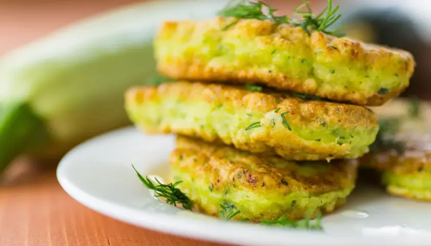 Frittelle di zucchine dorate e soffici, guarnite con aneto fresco, impilate su un piatto bianco. Sullo sfondo una zucchina intera.
