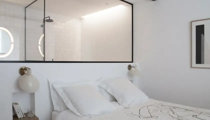 Stanza buia con letto bianco, una finestra tra due stanze rivela un bagno moderno con doccia e specchi luminosi.