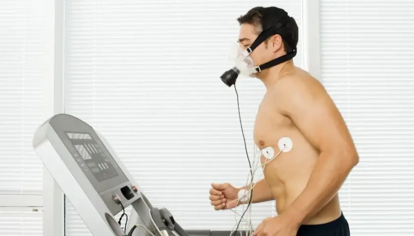 Mężczyzna w masce biegający na bieżni, podłączony do aparatury EKG. Wyniki testu VO2max w tabeli na ekranie bieżni.