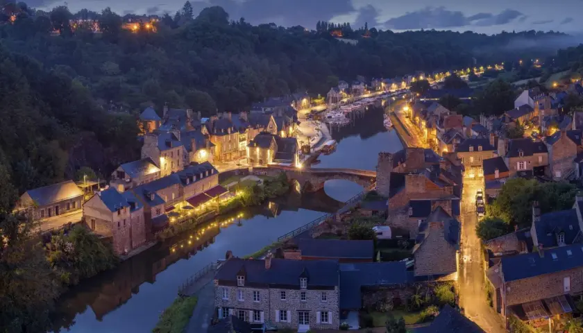 Widok na francuskie miasteczko nad rzeką o zmierzchu, z oświetlonymi budynkami i łodziami.