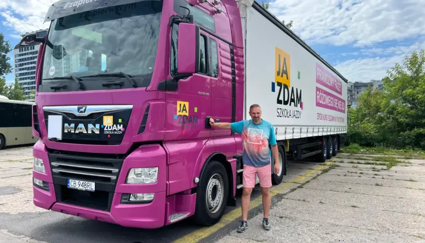 Mężczyzna stoi obok różowej ciężarówki MAN z naczepą. Na naczepie napis "JA ZDAM SZKOŁA JAZDY". To idealne miejsce, by zdobyć prawo jazdy c+e.