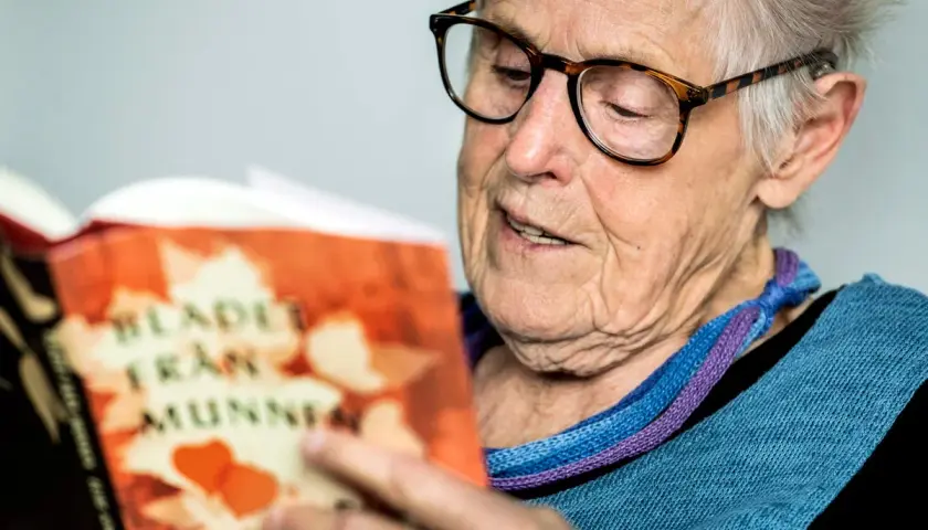 Äldre kvinna med glasögon läser en bok med titeln "Blad från munnen" av Elsie Johansson Thore.