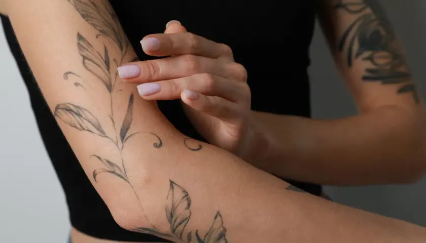 Delikatny tatuaż gałązki z liśćmi na przedramieniu. Dłonie z jasnym manicurem dotykają skóry, podkreślając, ile goi się tatuaż.