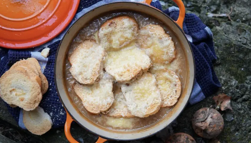 Zuppa di cipolle fumante in una casseruola arancione, con crostini dorati e formaggio fuso. Accanto, pane tostato e cipolle crude.