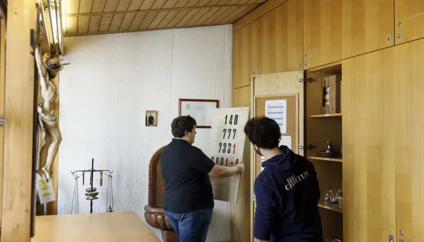 Zwei Männer im katholischen Priesterseminar hängen eine Liste mit Zahlen auf. Ein Kruzifix hängt an der Wand.