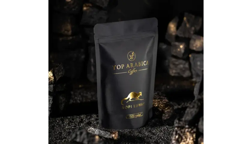 Czarna torebka z napisem "TOP ARABICA Coffee Kopi Luwak" i złotym rysunkiem zwierzęcia. To najdroższa kawa rozpuszczalna, otoczona węglem i złotymi drobinkami.