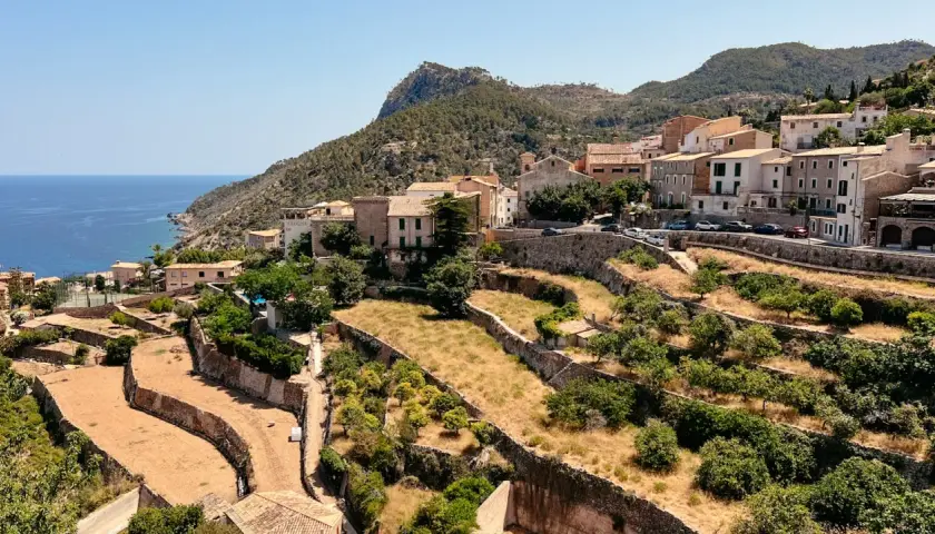 Terrassierte Hänge mit Bäumen und trockener Vegetation, die sich zum Meer hin erstrecken. Im Hintergrund liegt ein Dorf am Fuße des Tramuntana Gebirges.