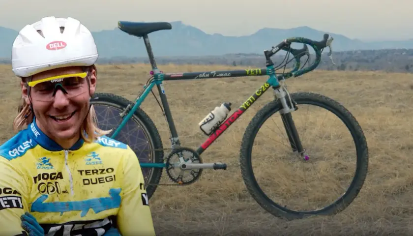 A smiling cyclist in a yellow jersey stands beside a vintage YETI C-26 bike, hinting at where YETI bikes are made.