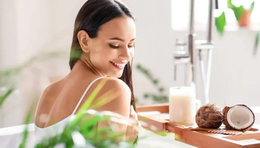 Mujer sonriente en la bañera con cocos y un frasco de aceite de coco, mostrando para qué sirve el aceite de coco para el cabello.