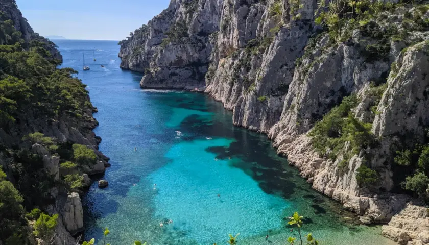 Aguas turquesas y cristalinas en el **calanque d'en Vau**, rodeado de imponentes acantilados rocosos y vegetación. Gente nadando y barcos a lo lejos.