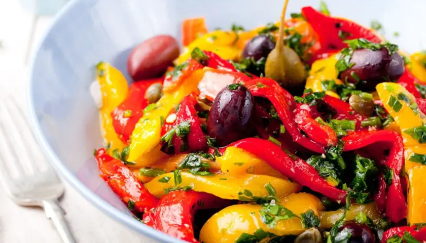 Insalata colorata di peperoni in agrodolce, con olive nere, capperi e prezzemolo fresco, servita in una ciotola blu.