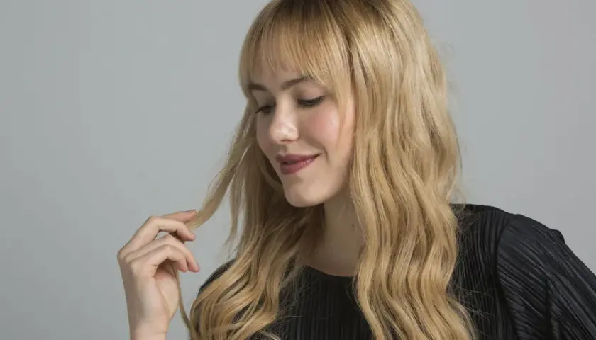 Mujer con cabello rubio ondulado y flequillo, tocando un mechón. El matizar el pelo realza su brillo y tono natural.